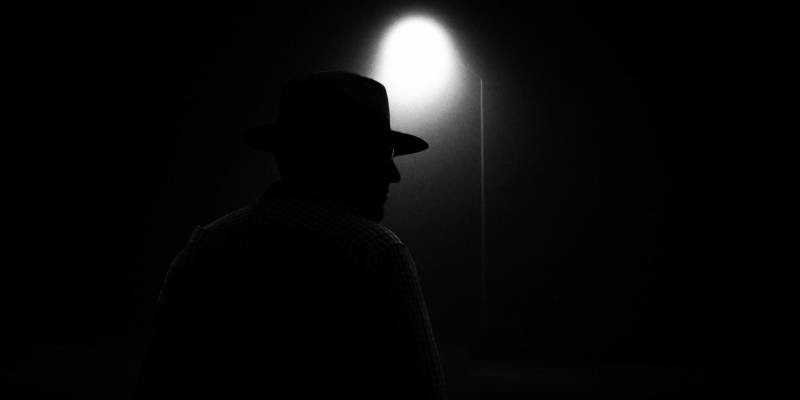 The silhouette of a man in a wide brimmed hat lit under the glow of a distant street lamp.