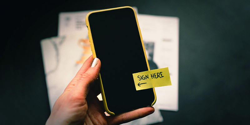 Ac lose of of a hand holding an iPhone. The iPhone's display is switched off and a post-it note is attached with the words sign here written on it.