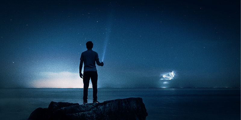 A person standing on a rock at waters edge at night their flashlight beam piercing the darkness.